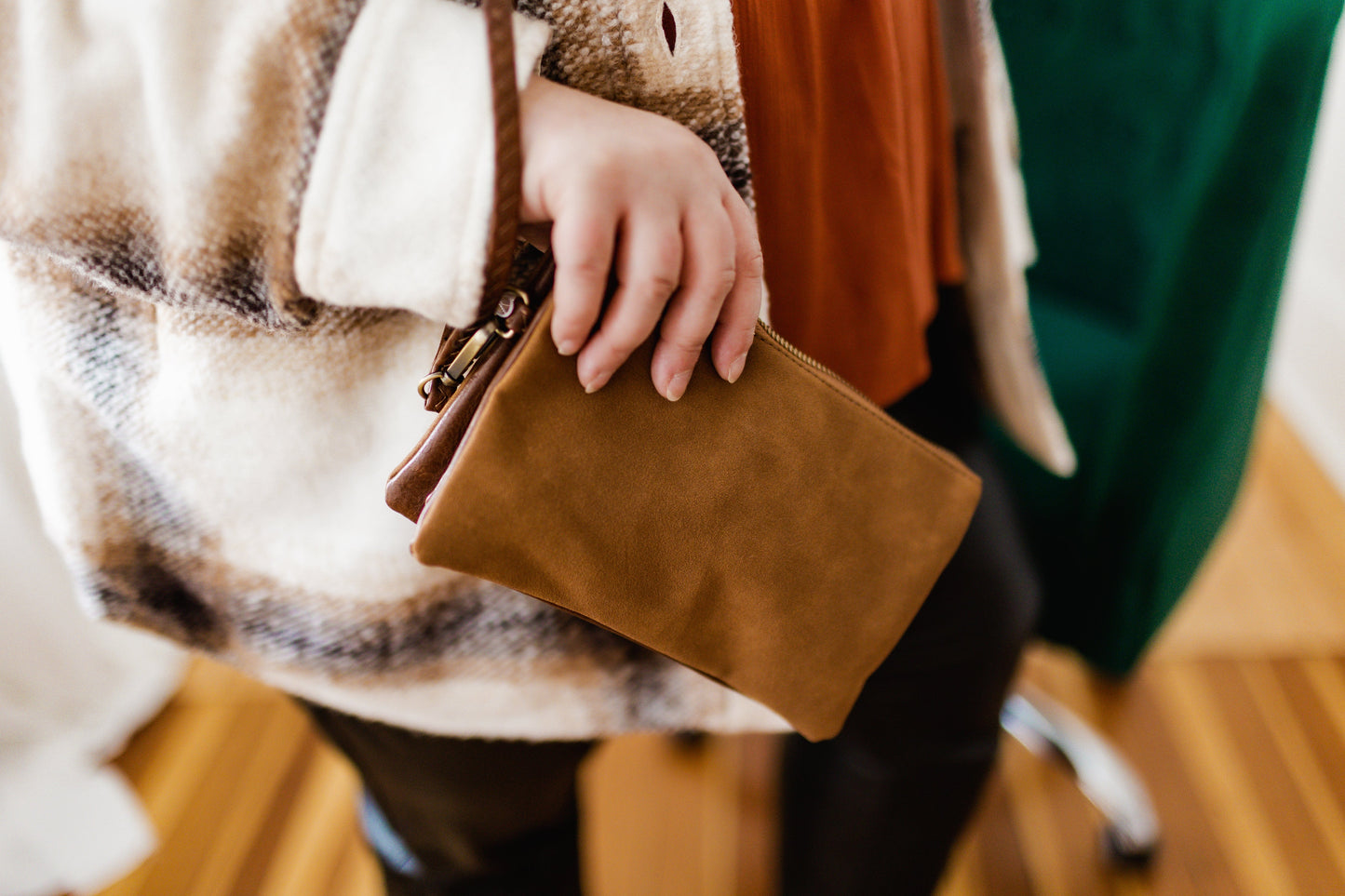 Person holding a brown faux suede clutch with a detachable strap, wearing a plaid jacket and standing on a wooden floor.