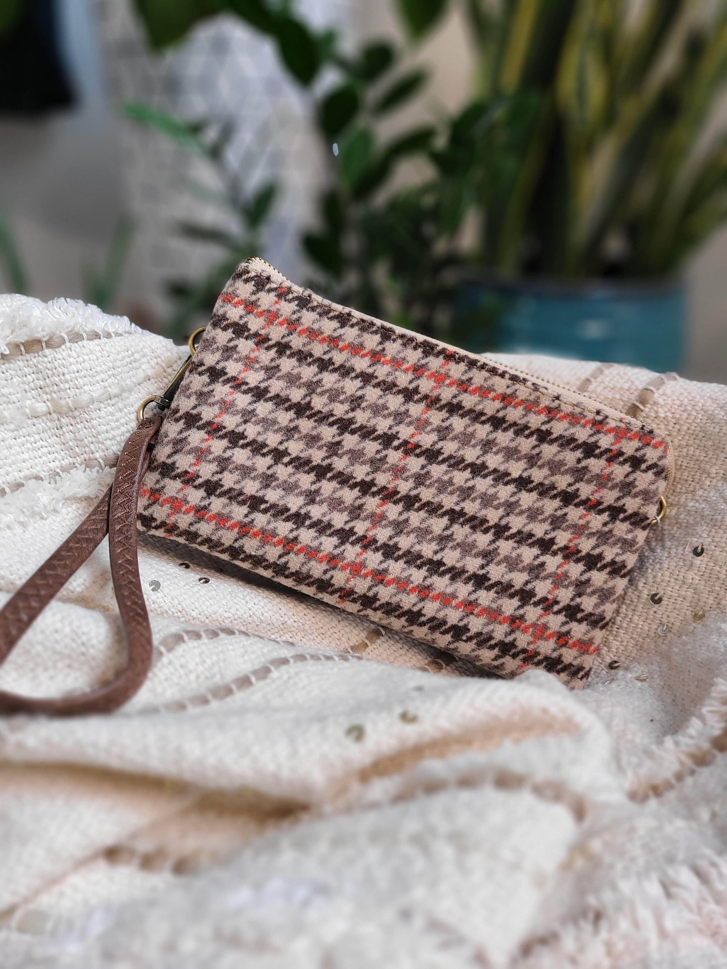A chic houndstooth-patterned clutch wallet, featuring a faux leather design and a detachable wrist strap, rests elegantly on a textured white fabric backdrop.