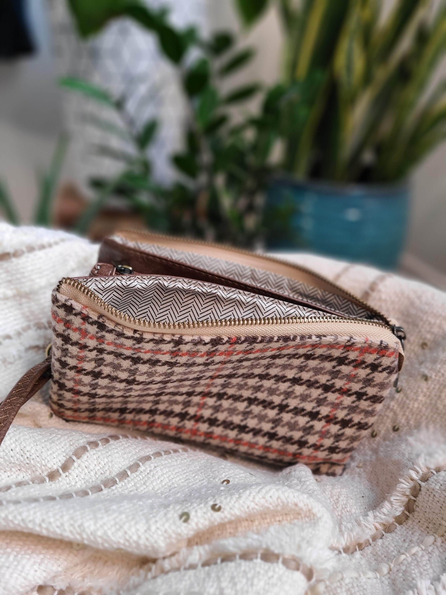 A plaid-patterned clutch with a zipper and detachable strap sits on a textured fabric, with plants visible in the blurred background.
