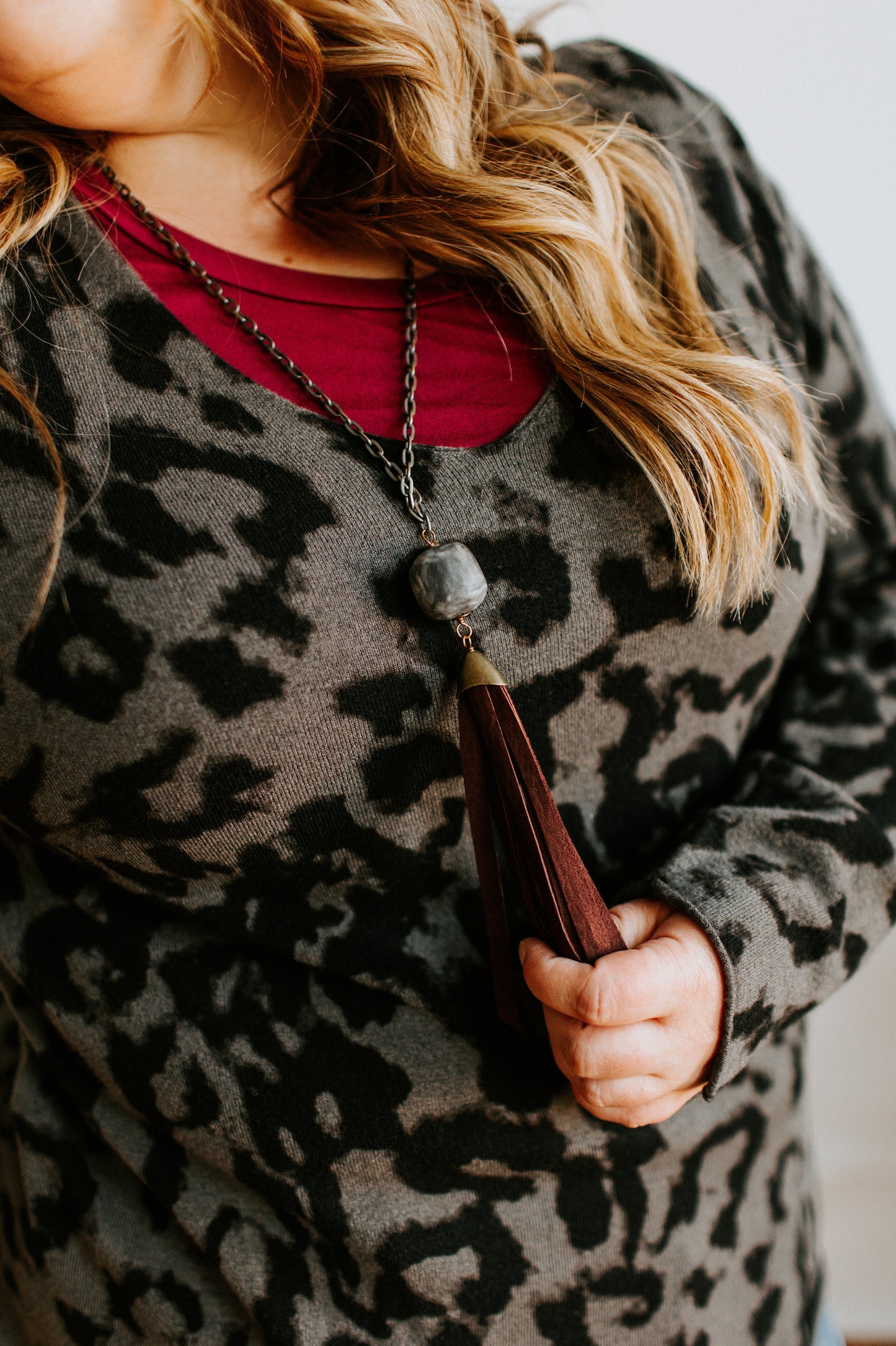 Close-up of a person in a leopard print top, showcasing a long necklace with a faux leather tassel, standing with one hand holding the tassel.