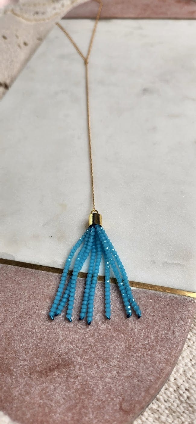 A gold necklace with a thin chain and a turquoise beaded tassel pendant, made in the USA, sits elegantly on a marble and pink surface.