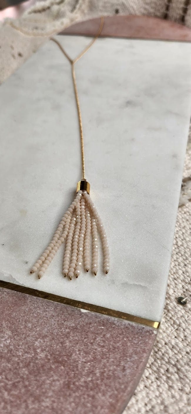 A gold beaded tassel necklace with cream-colored beads meticulously crafted and made in the USA, beautifully displayed on a rectangular marble and wood surface.