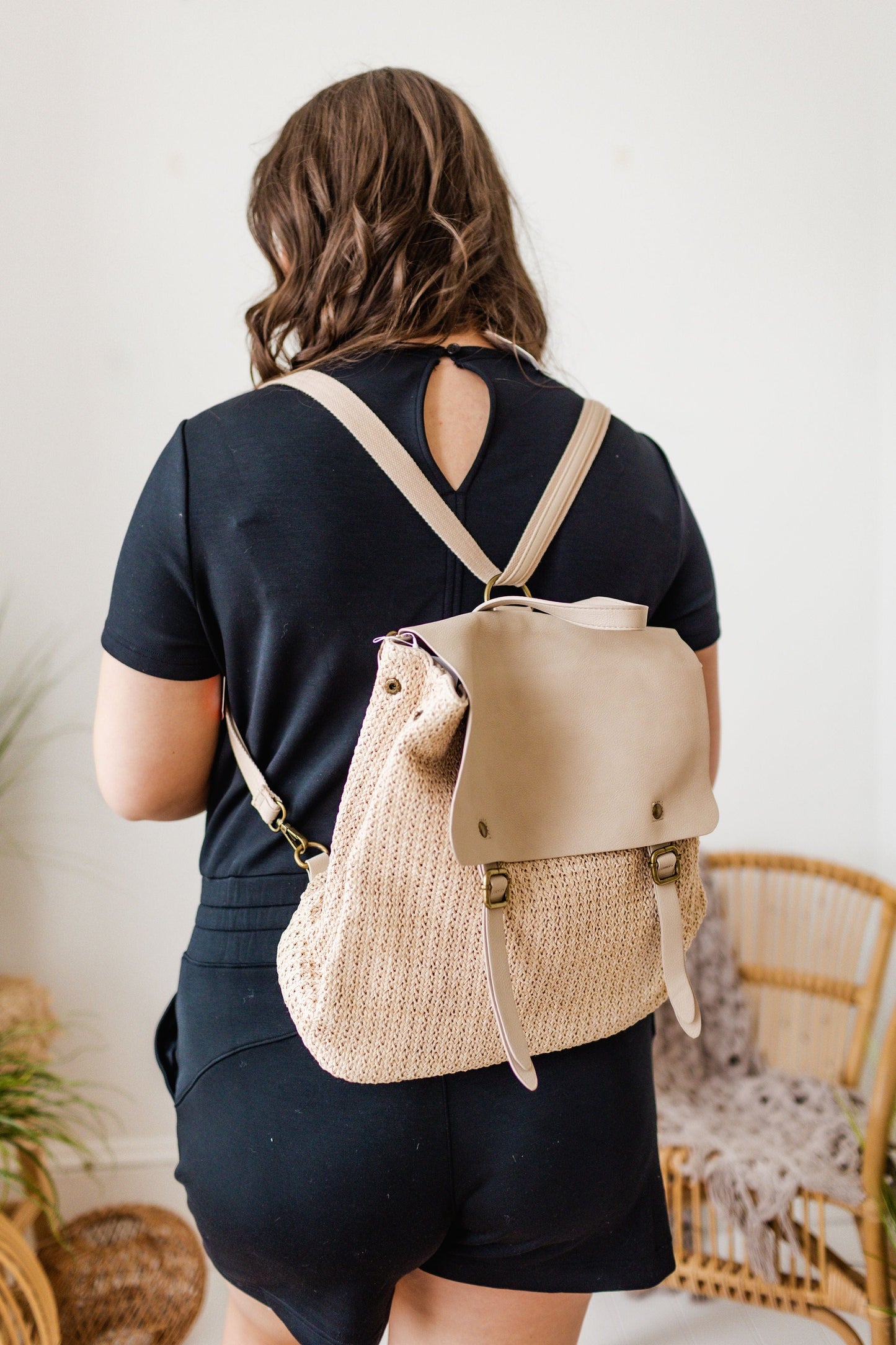 A person with long hair wearing a black outfit is seen from the back, carrying a beige woven summer bag made with vegan leather straps.