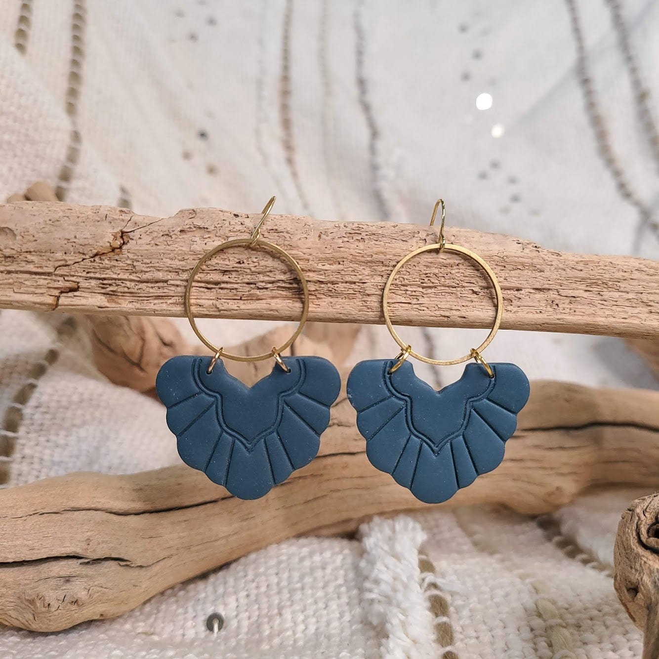 Blue scallop-shaped polymer clay earrings with brass hoops hang elegantly on a piece of driftwood. The background showcases textured fabric and additional driftwood, adding an organic feel to the scene.