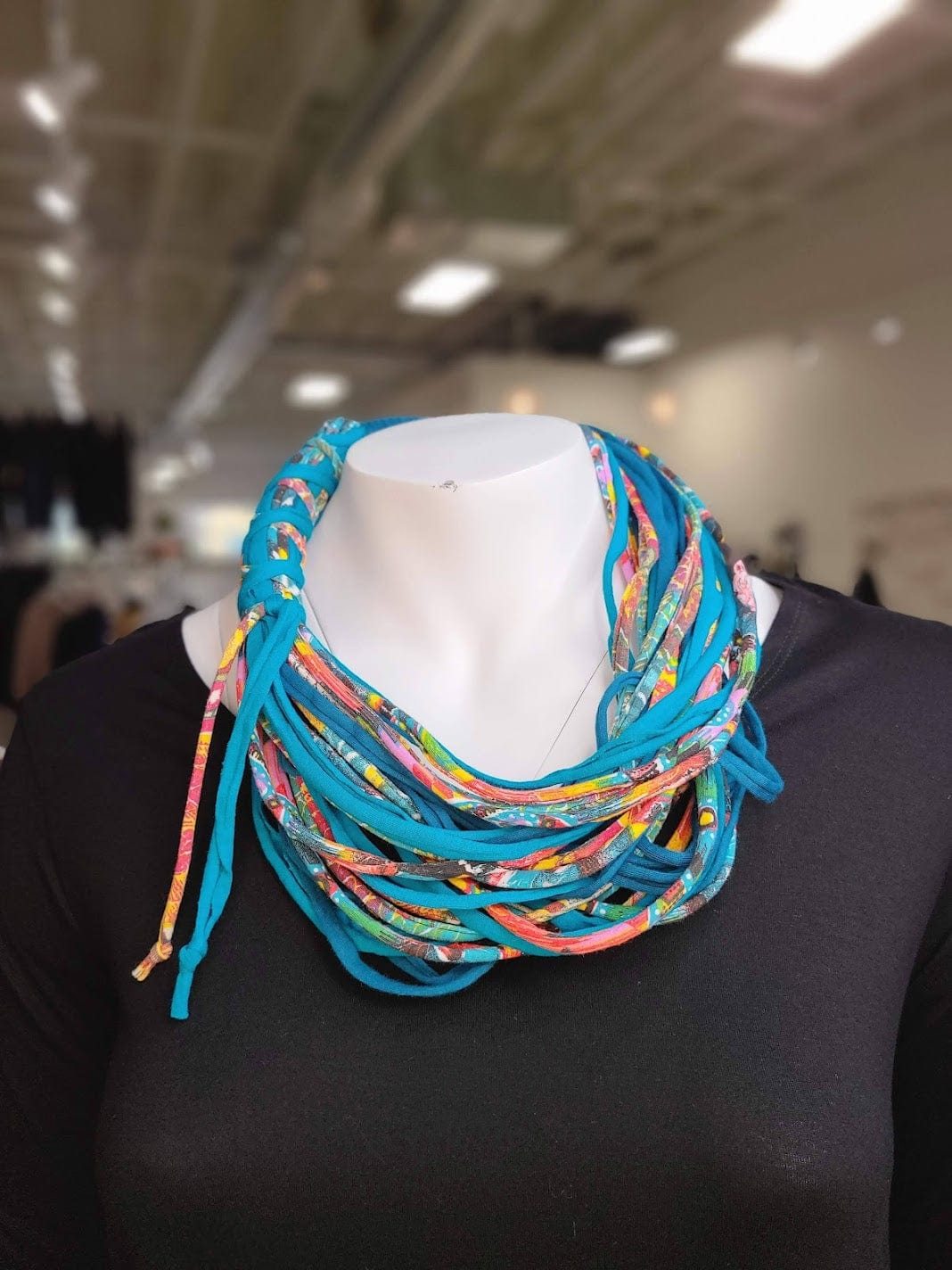 A mannequin wearing a black top is adorned with a colorful, multi-layered fabric necklace crafted from textile remnants in a well-lit indoor setting.