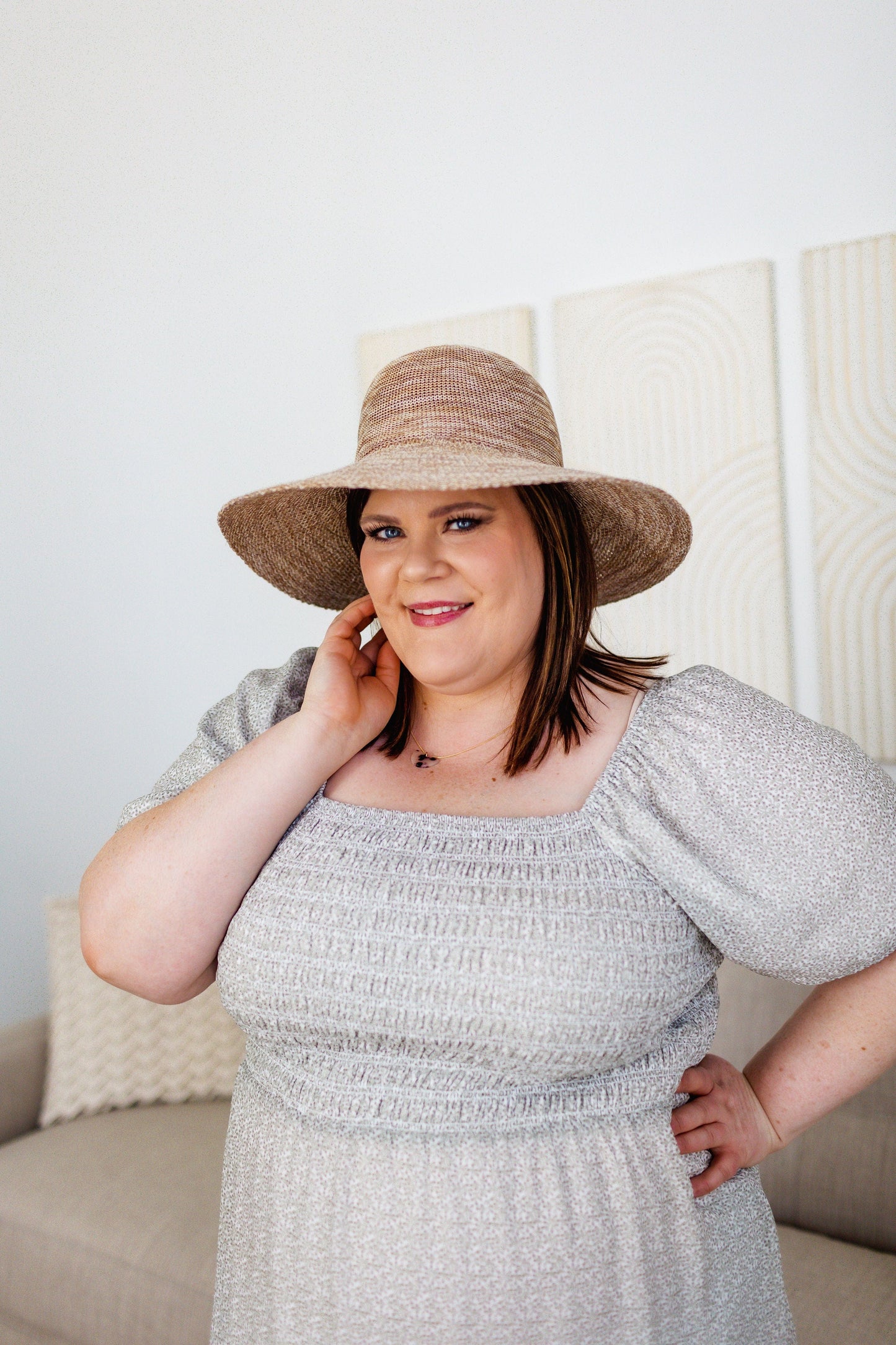 A woman wearing a wide-brim straw hat and a light-colored dress stands indoors, smiling with one hand on her hip and the other touching her face, exuding effortless elegance and sun protection charm.