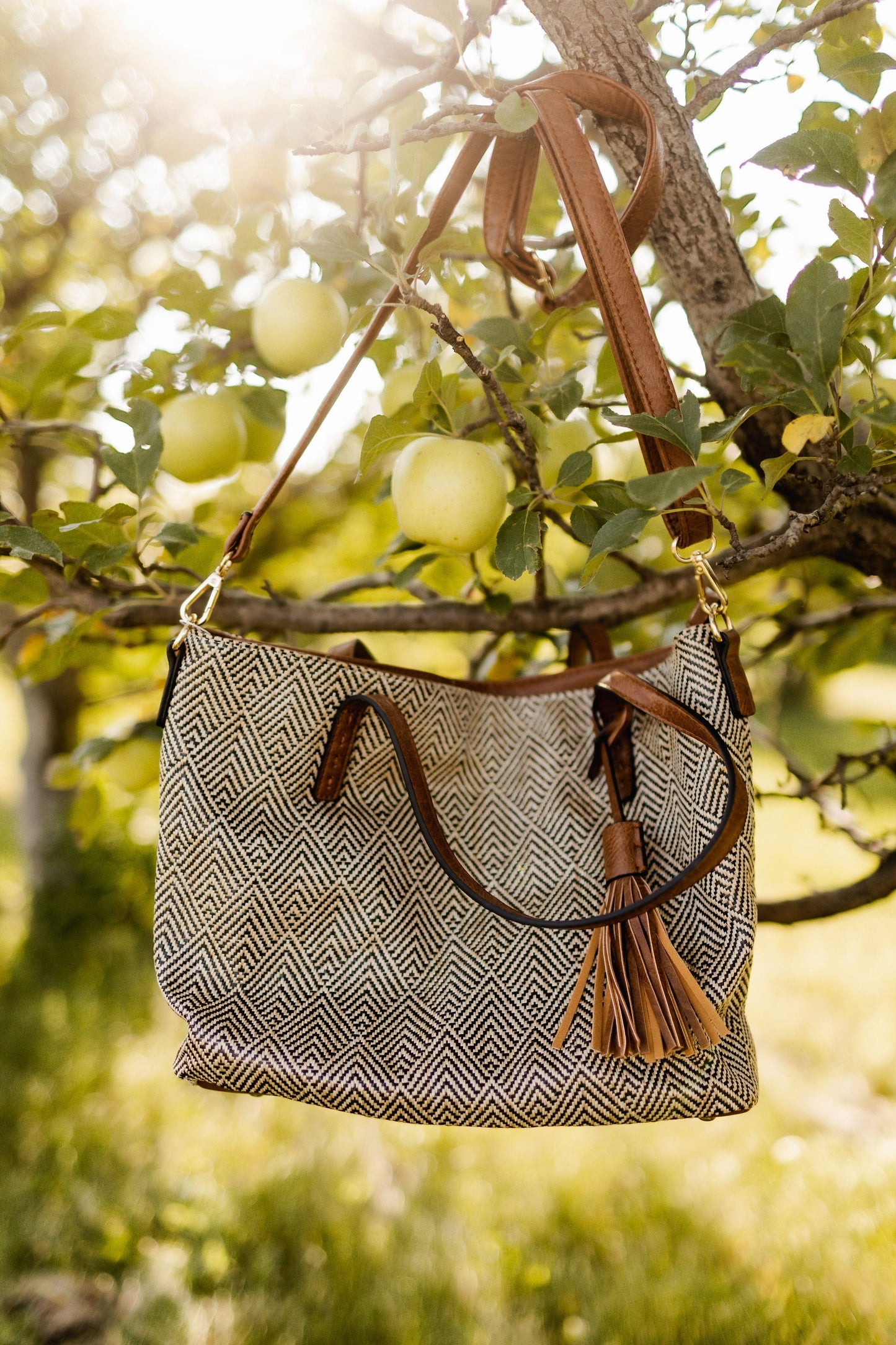 A patterned woven hobo bag with a tassel hangs on a tree branch among green apples in sunlight.