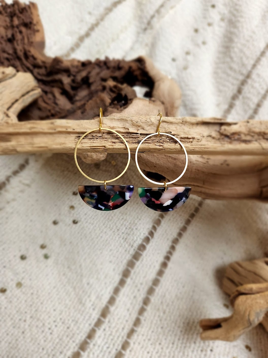Gold hoop earrings with semicircular, multicolored pendants crafted from Lucite are elegantly displayed on driftwood against a textured fabric background. Handmade in the USA, these unique pieces come with free shipping for your added convenience.