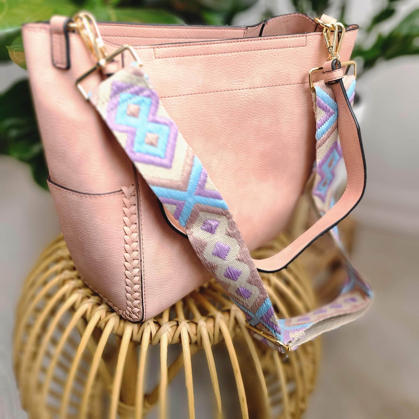 A pink vegan faux leather handbag with a patterned strap rests elegantly on a woven stool, offering the versatility of a detachable shoulder strap.