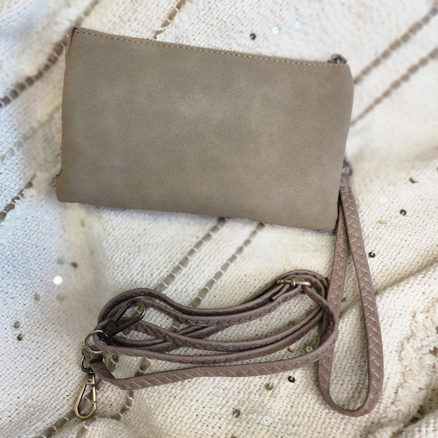 A taupe color clutch bag with a matching detachable strap and metal clasp sits elegantly on a white textured fabric.