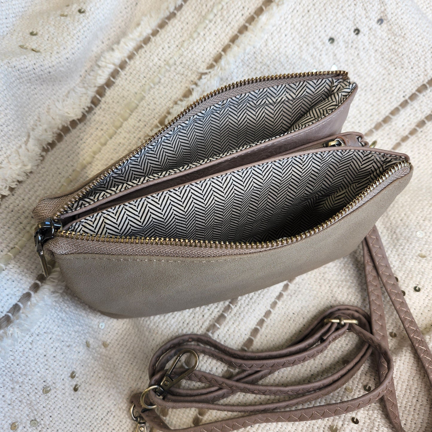 A taupe-colored crossbody bag with two zippered compartments, chevron-patterned lining, and an adjustable strap lies open on a textured surface.