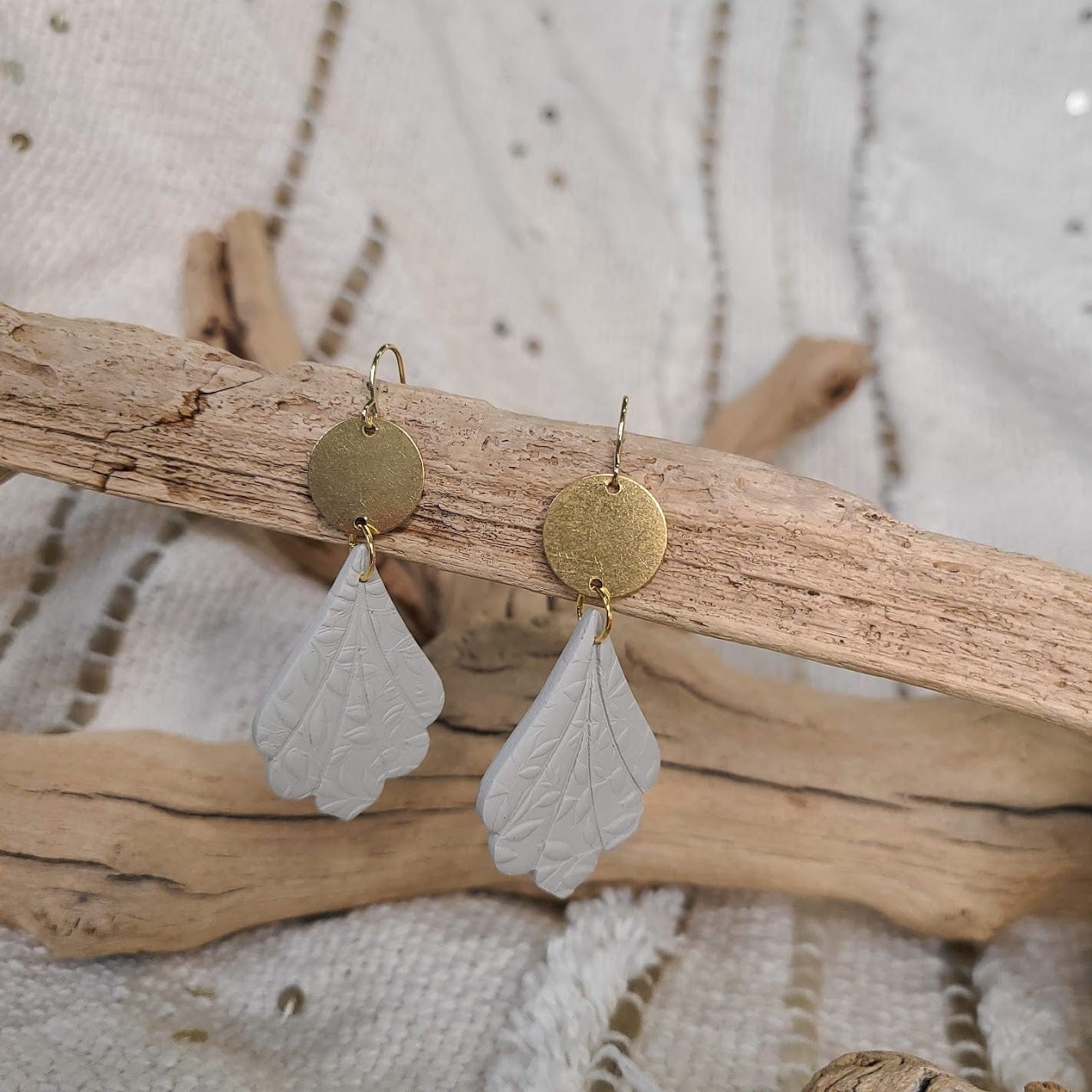 Gold discs with white leaf-shaped polymer clay earrings hang gracefully on a wooden branch. These stunning pieces are finished with gold plated hooks, adding a touch of elegance to any outfit.