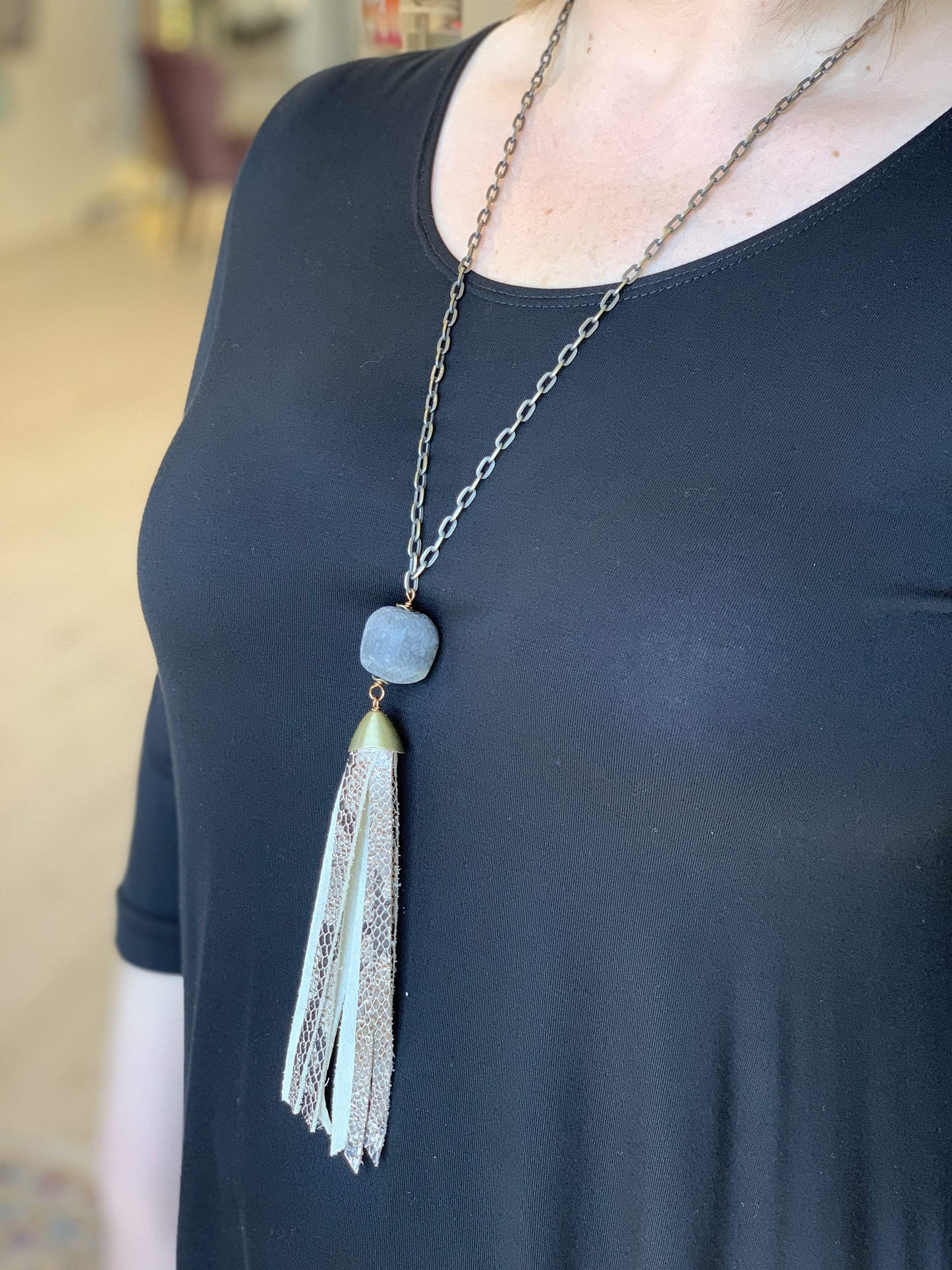 A woman wearing a black top and a long necklace featuring a square stone and faux leather tassel pendant.