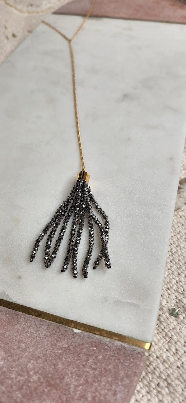 A stunning gold necklace with a fine chain showcases a beaded tassel pendant crafted from multiple strands of small, dark beads. Displayed elegantly on a marble surface, this piece proudly bears the mark of quality as it is made in the USA.