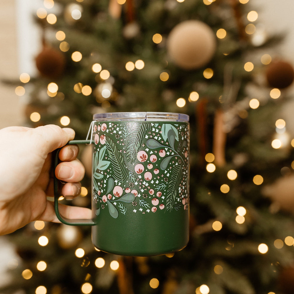 A hand holds a green, triple-walled mug with a festive design, brimming with holiday cheer, in front of a glowing Christmas tree adorned with golden lights.