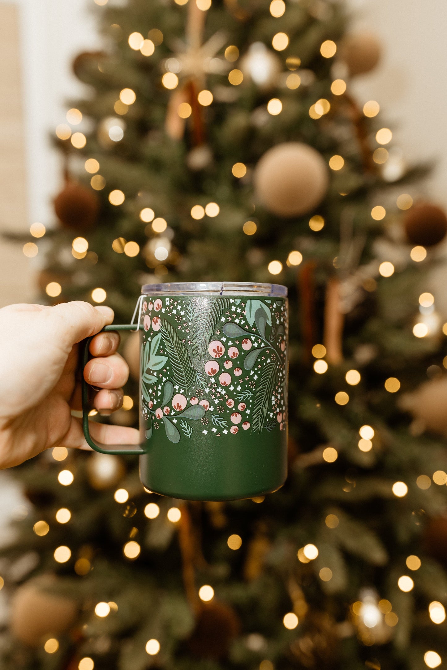 A hand holds a green, triple-walled mug with a festive design, brimming with holiday cheer, in front of a glowing Christmas tree adorned with golden lights.