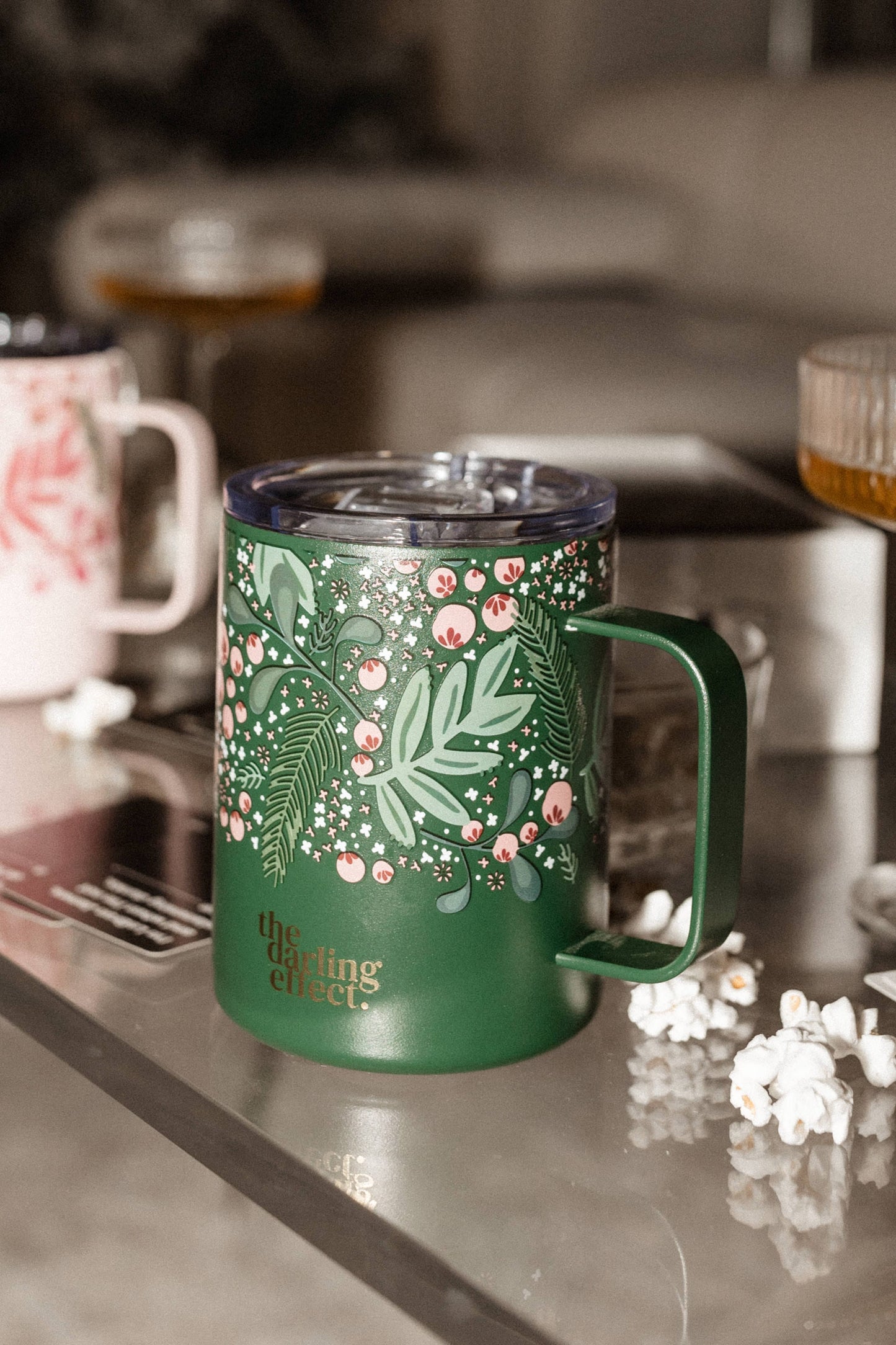 Green mug with floral design and the text "the darling effect" on a table, surrounded by popcorn and another triple-walled insulated mug in the background.