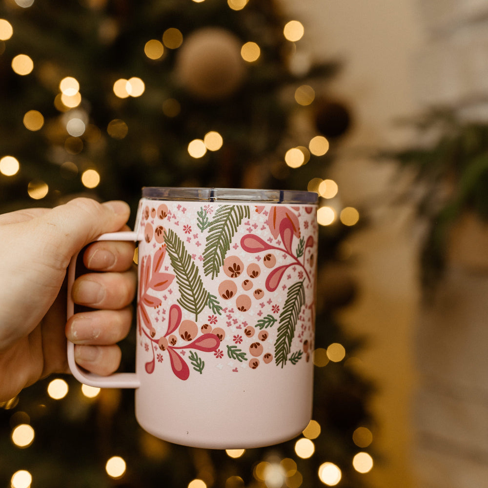 A hand holds a floral-patterned, BPA-free mug with triple-walled insulation in front of a blurred, illuminated Christmas tree.
