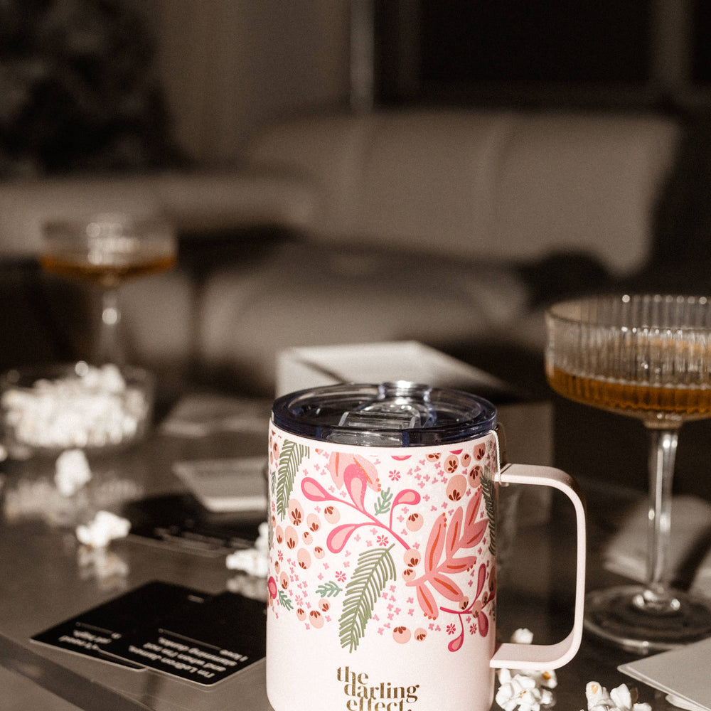 A BPA-free floral mug sits on a glass table, surrounded by playing cards and popcorn, with two stemmed glasses filled with a beverage in the background. The mug's triple-walled insulation keeps your drink perfectly warm amidst the lively setting.