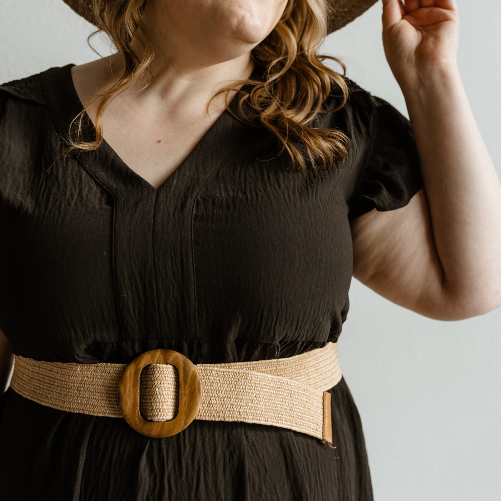 Woman wearing a black dress with a wide belt and a straw hat against a neutral background