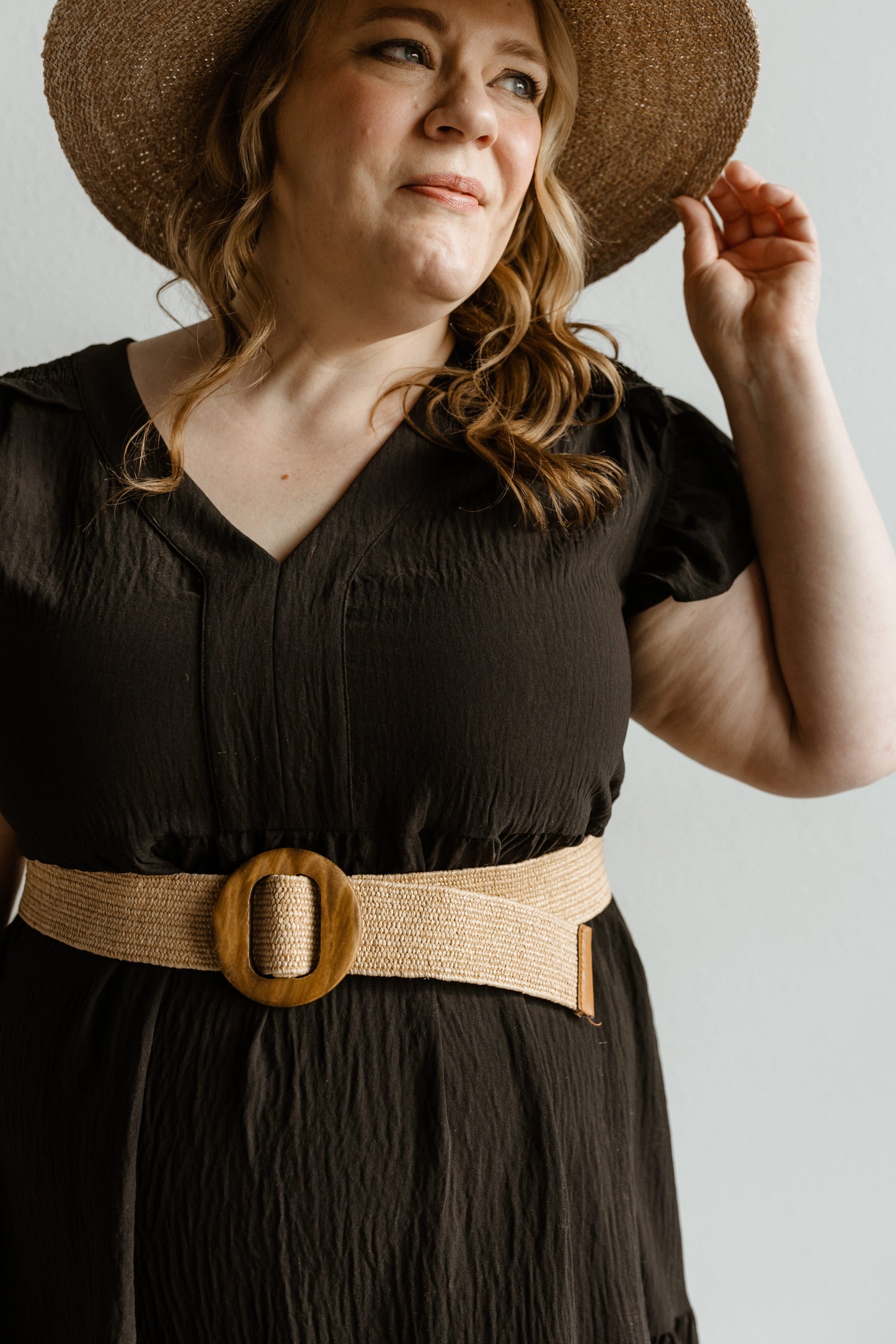 Woman wearing a black dress with a wide belt and a straw hat against a neutral background