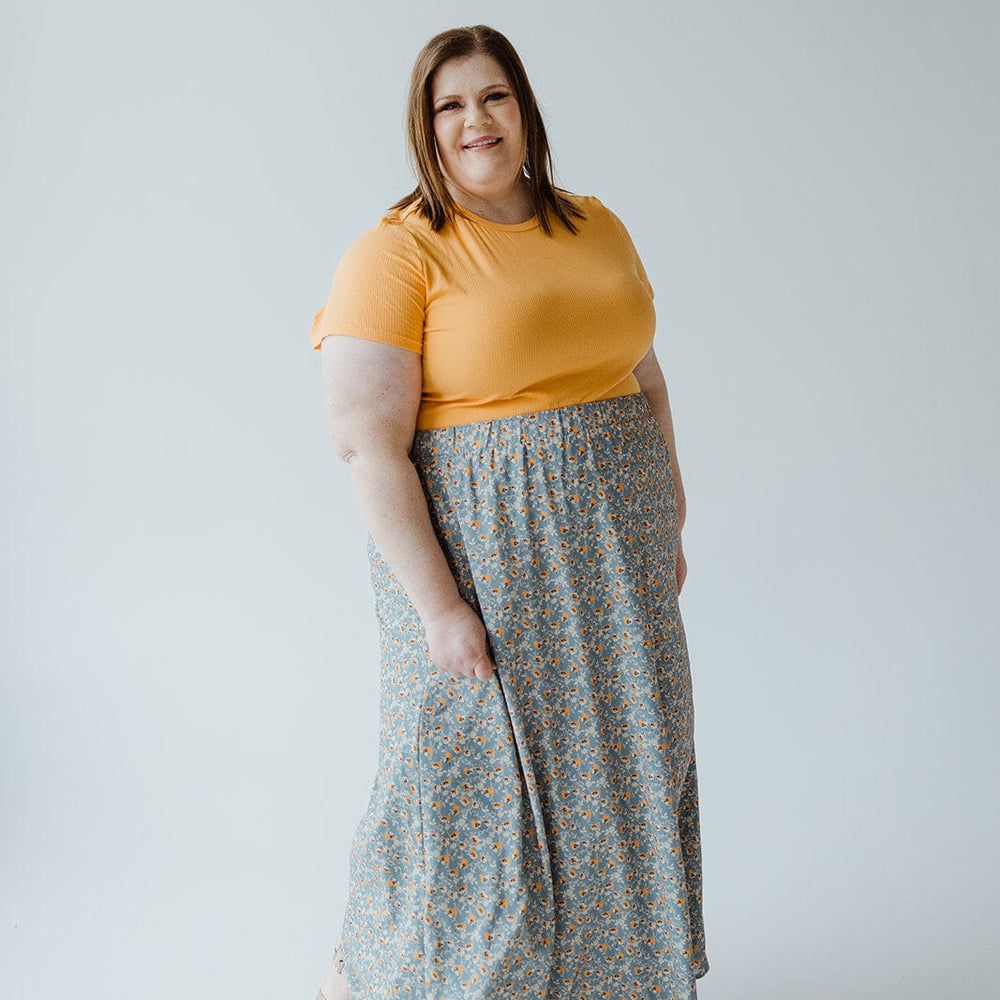 A woman with shoulder-length hair stands against a plain background, wearing a yellow short-sleeve shirt and an A-LINE MAXI SKIRT by Love Marlow with a floral pattern, holding a piece of the skirt with her right hand.