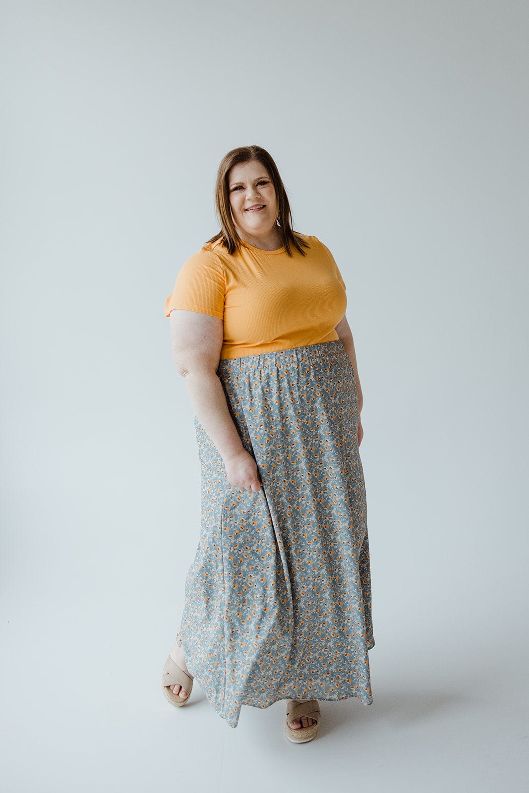 A woman with shoulder-length hair stands against a plain background, wearing a yellow short-sleeve shirt and an A-LINE MAXI SKIRT by Love Marlow with a floral pattern, holding a piece of the skirt with her right hand.