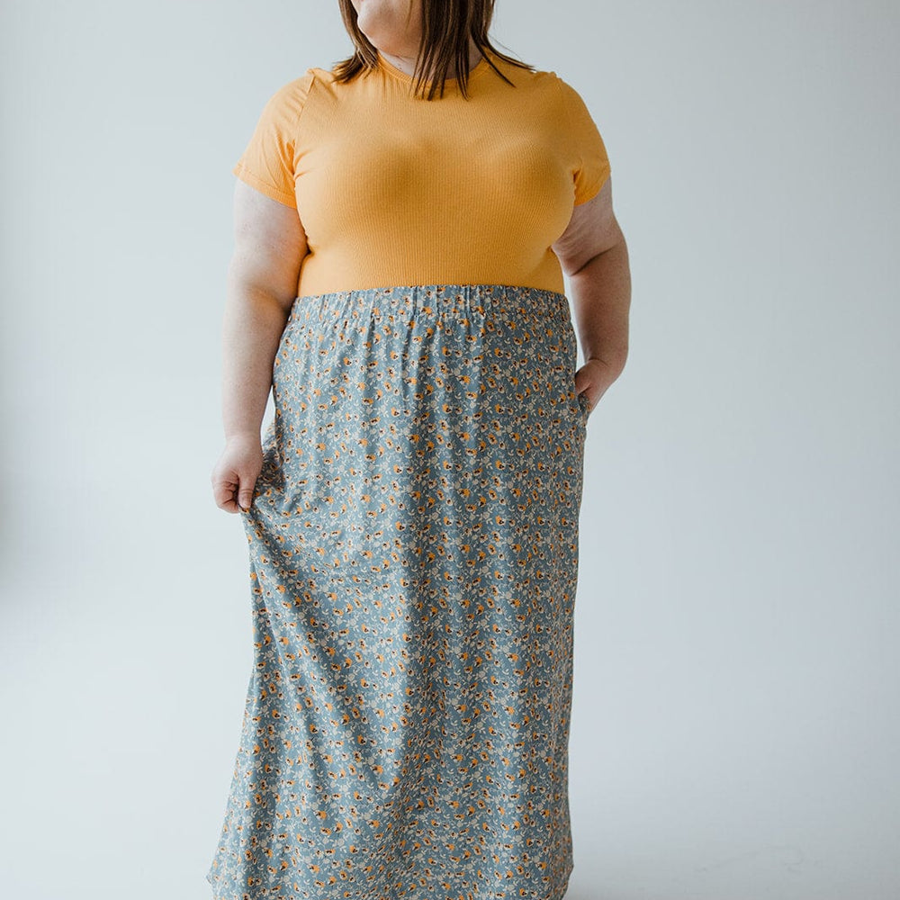 A person in a yellow shirt and long Love Marlow A-LINE MAXI SKIRT with a floral pattern stands against a plain background, holding the skirt with one hand and looking to the side.