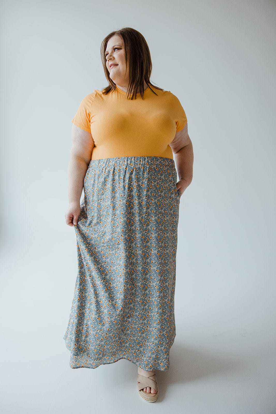 A person in a yellow shirt and long Love Marlow A-LINE MAXI SKIRT with a floral pattern stands against a plain background, holding the skirt with one hand and looking to the side.