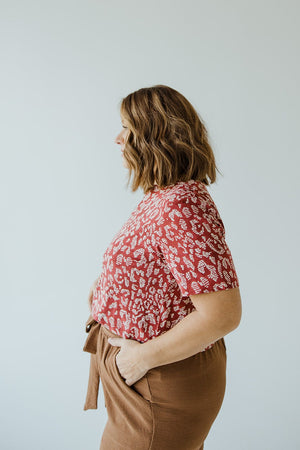 A person with shoulder-length hair, wearing a Love Marlow ABSTRACT ANIMAL PRINT SHORT SLEEVE TEE and brown pants, standing in profile against a plain light background.