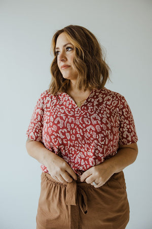 A person with shoulder-length hair stands against a plain background, wearing a Love Marlow ABSTRACT ANIMAL PRINT SHORT SLEEVE TEE and brown pants with a tie waist.