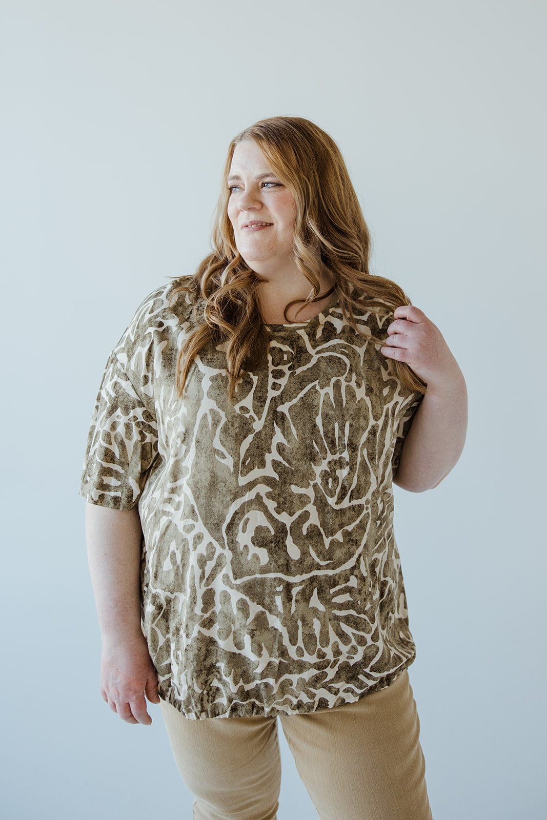 A person with long hair and a relaxed expression wears an ABSTRACT PRINT BLOUSE WITH TIE DETAIL IN FOREST MOSS by Love Marlow, standing against a plain background.