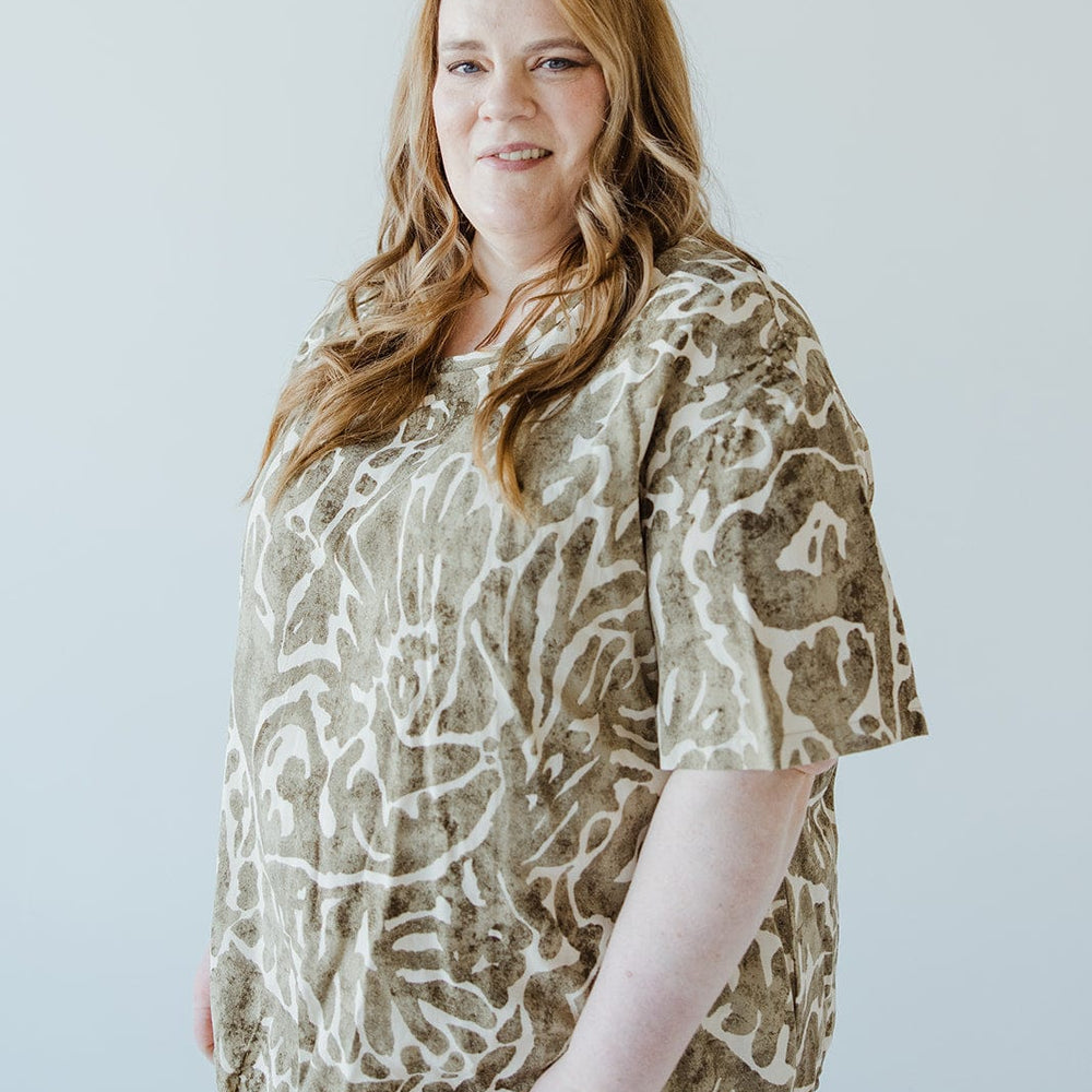 A woman with long hair is standing and wearing a Love Marlow ABSTRACT PRINT BLOUSE WITH TIE DETAIL IN FOREST MOSS with light-colored pants. She is looking at the camera and smiling slightly. The background is plain and light-colored.