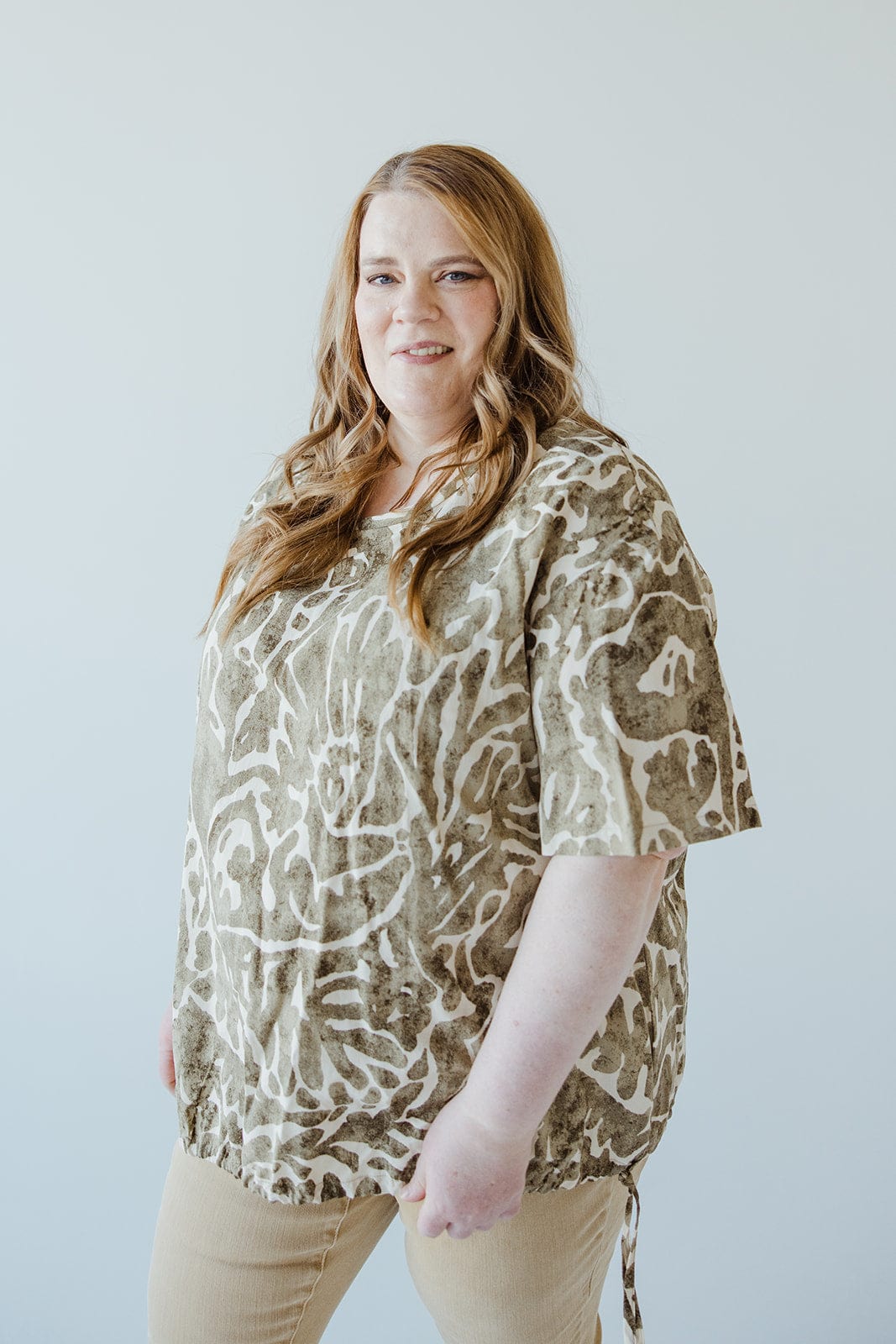 A woman with long hair is standing and wearing a Love Marlow ABSTRACT PRINT BLOUSE WITH TIE DETAIL IN FOREST MOSS with light-colored pants. She is looking at the camera and smiling slightly. The background is plain and light-colored.