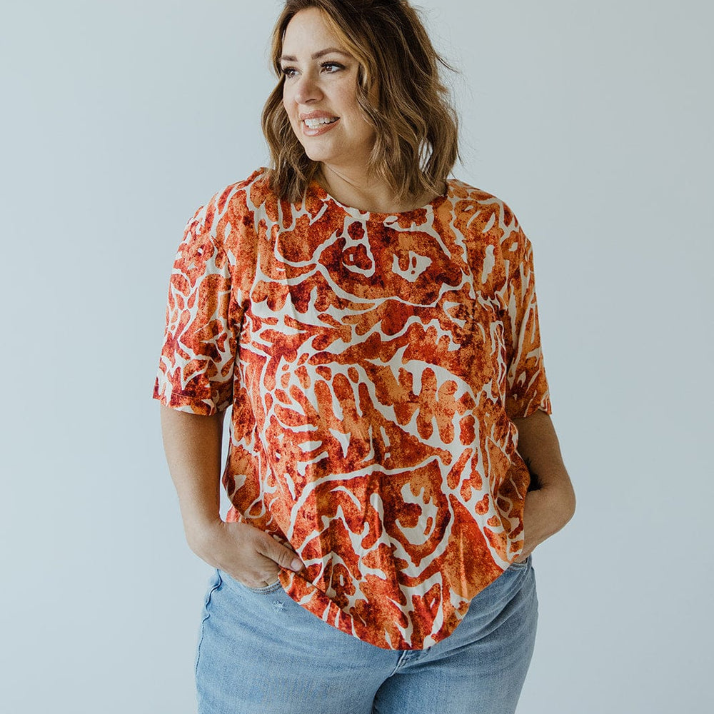 A woman with wavy hair, dressed in the luxurious "ABSTRACT PRINT BLOUSE WITH TIE DETAIL IN HAWAIIAN HIBISCUS" by Love Marlow, paired with blue jeans, stands with her hands in her pockets against a plain background.