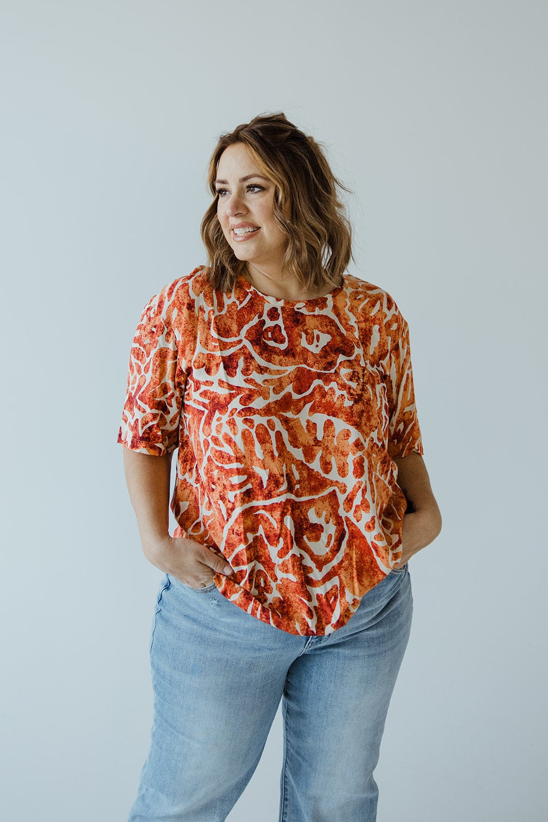 A woman with wavy hair, dressed in the luxurious "ABSTRACT PRINT BLOUSE WITH TIE DETAIL IN HAWAIIAN HIBISCUS" by Love Marlow, paired with blue jeans, stands with her hands in her pockets against a plain background.