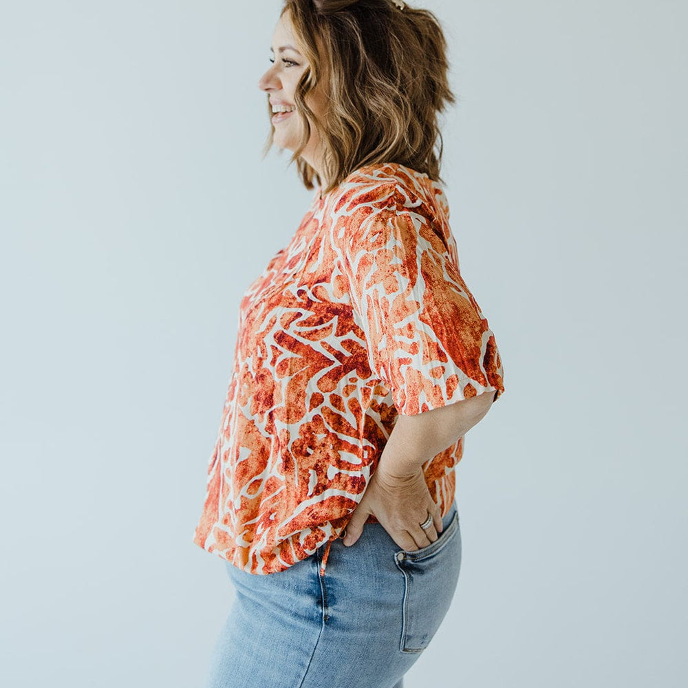 A woman with shoulder-length hair stands in profile, wearing a luxurious Love Marlow Abstract Print Blouse with Tie Detail in Hawaiian Hibiscus and blue jeans against a plain background.