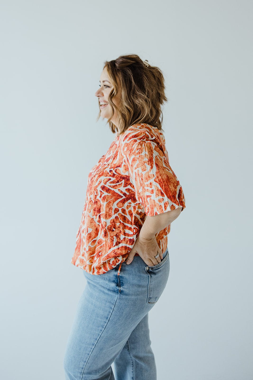 A woman with shoulder-length hair stands in profile, wearing a luxurious Love Marlow Abstract Print Blouse with Tie Detail in Hawaiian Hibiscus and blue jeans against a plain background.