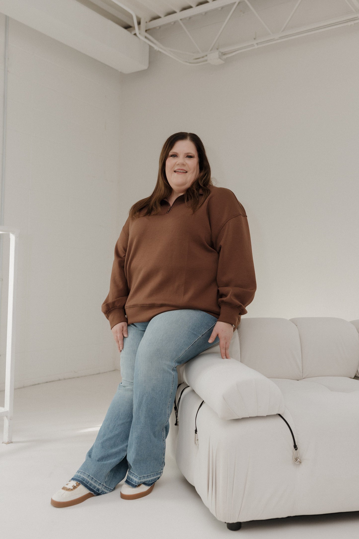 Person wearing a brown sweater and blue jeans sitting on a white couch in a minimalistic room.