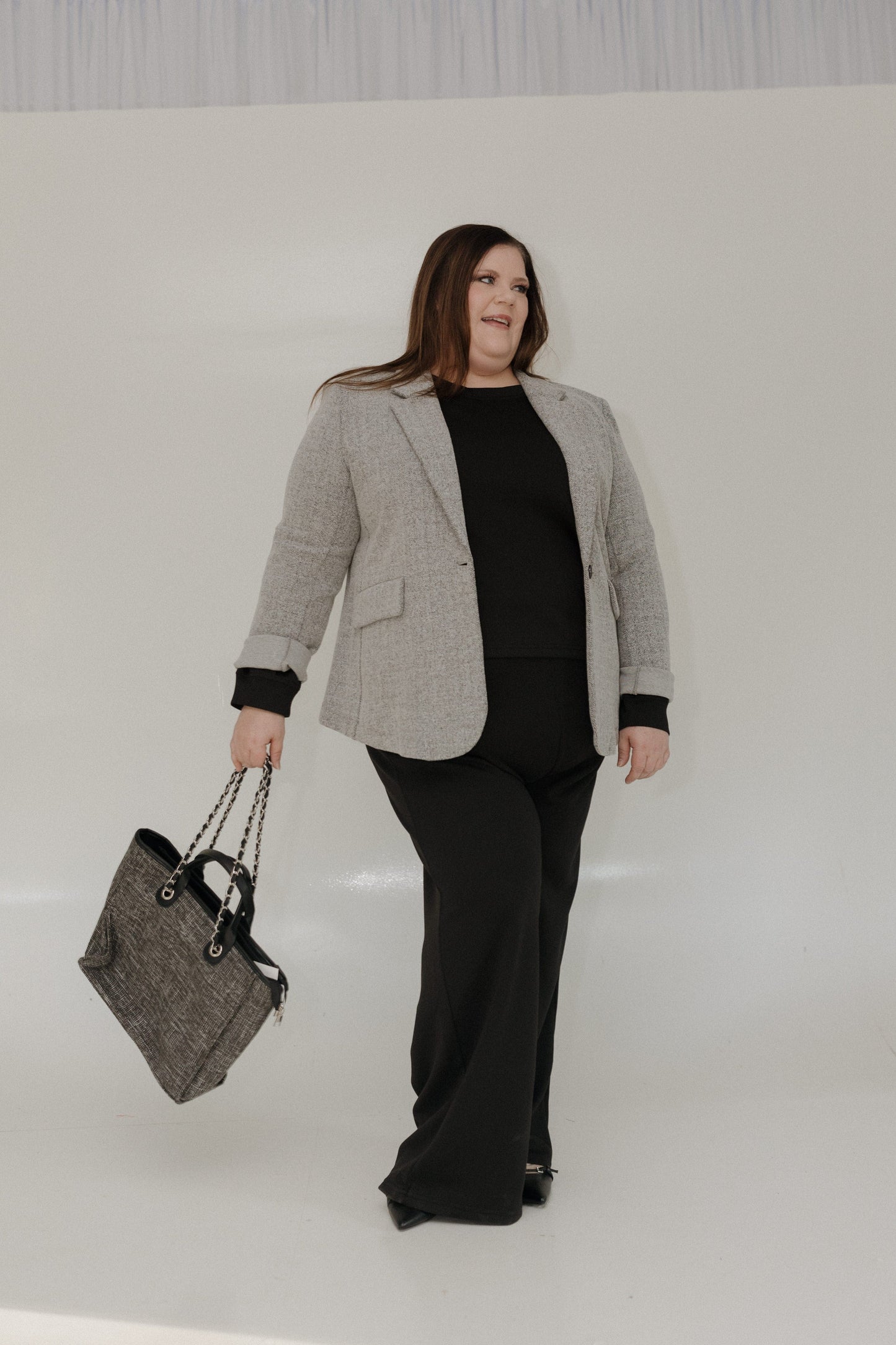 Woman in a gray blazer and black pants holding a gray bag against a white background