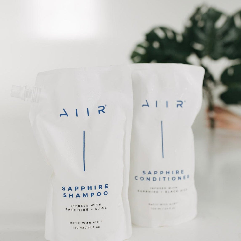 Two bags of AIIR Sapphire Shampoo and Conditioner on a white surface with a blurred plant in the background.