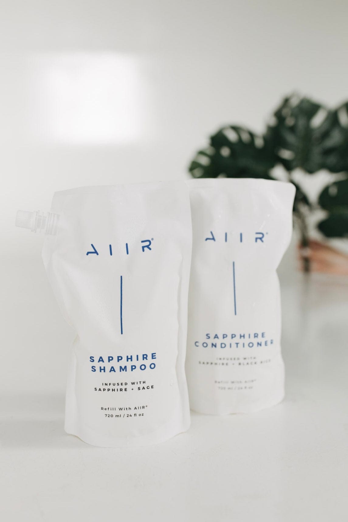 Two bags of AIIR Sapphire Shampoo and Conditioner on a white surface with a blurred plant in the background.