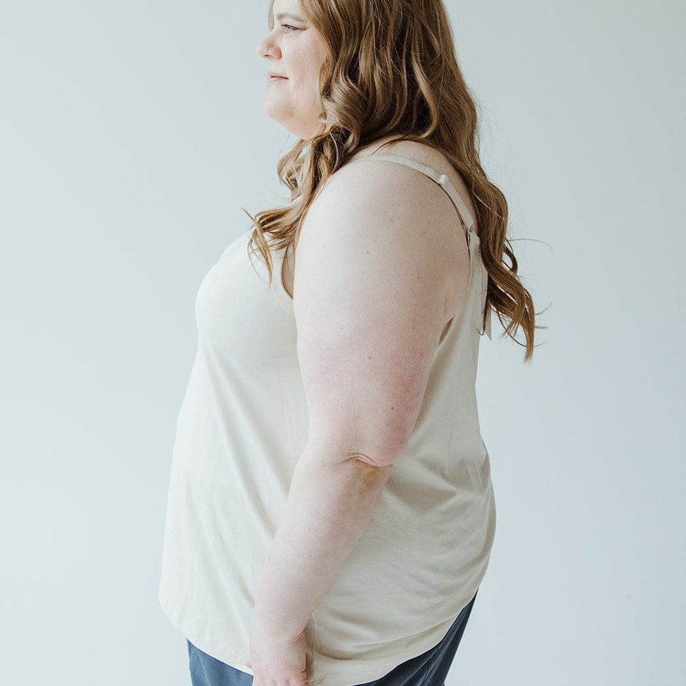 A woman with long brown hair, dressed in Love Marlow's BASIC LAYERING CAMI IN LIGHT BEIGE paired with blue pants, stands in a side profile view against a plain background.