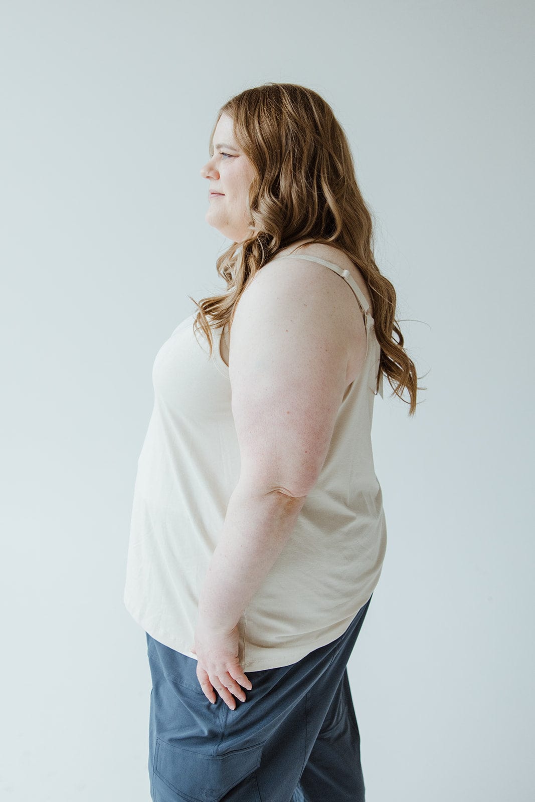 A woman with long brown hair, dressed in Love Marlow's BASIC LAYERING CAMI IN LIGHT BEIGE paired with blue pants, stands in a side profile view against a plain background.