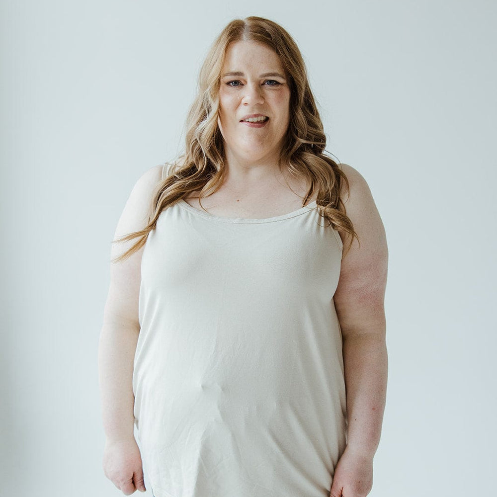 Against a plain backdrop, a person with long hair is dressed in the Love Marlow BASIC LAYERING CAMI IN LIGHT BEIGE paired with dark pants.