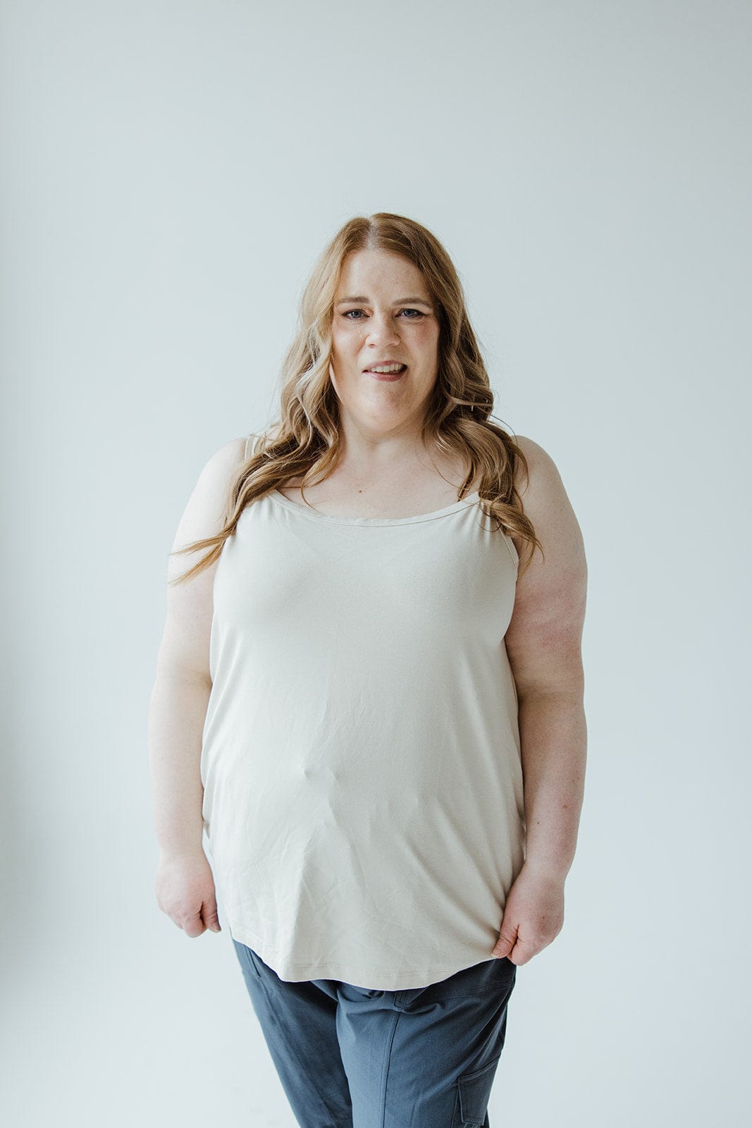 Against a plain backdrop, a person with long hair is dressed in the Love Marlow BASIC LAYERING CAMI IN LIGHT BEIGE paired with dark pants.