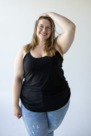 A person wearing glasses, the BASIC RACERBACK CAMI IN BLACK by Love Marlow, and jeans smiles while standing against a plain background, with one hand on their head.