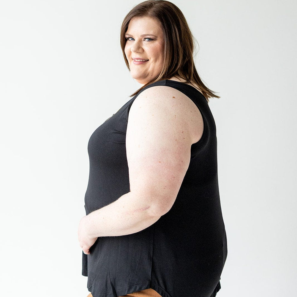 A woman with shoulder-length brown hair smiles while posing sideways. She is wearing a BASIC ROUND NECK TUNIC TANK IN BLACK by Love Marlow and brown pants, standing against a plain white background.