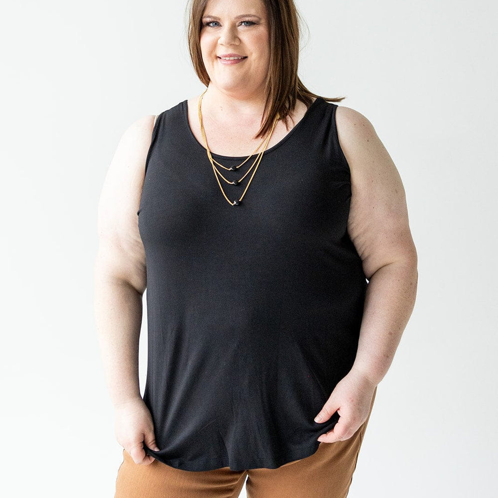 Person with brown hair, wearing a Love Marlow BASIC ROUND NECK TUNIC TANK IN BLACK featuring wide tank straps and a high-low rounded hem, along with brown pants. They are standing against a plain white background and smiling at the camera.