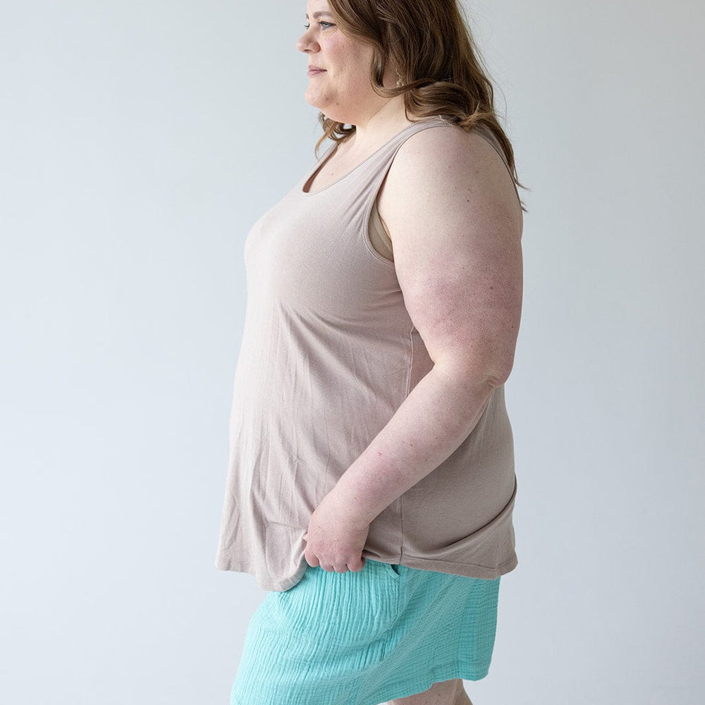 A person with long hair, wearing a chic and comfy BASIC ROUND NECK TUNIC TANK IN FROSTED TOFFEE by Love Marlow and turquoise shorts, stands against a plain white background, looking to the side with a hand in their pocket.